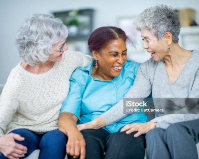 Three (3) diverse senior woman laugh while embracing each other. They are happy and exuding joy.