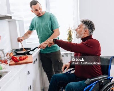 A mature Caucasian man in a wheelchair is instructing a young Caucasian man, who's volunteering to help him in the kitchen.