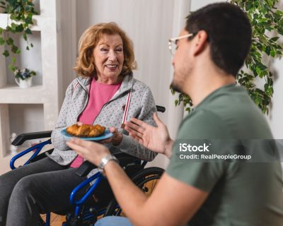 A man offers a plate with a croissant to an elderly woman in a wheelchair who smiles warmly at him in a cozy indoor setting with plants in the background.